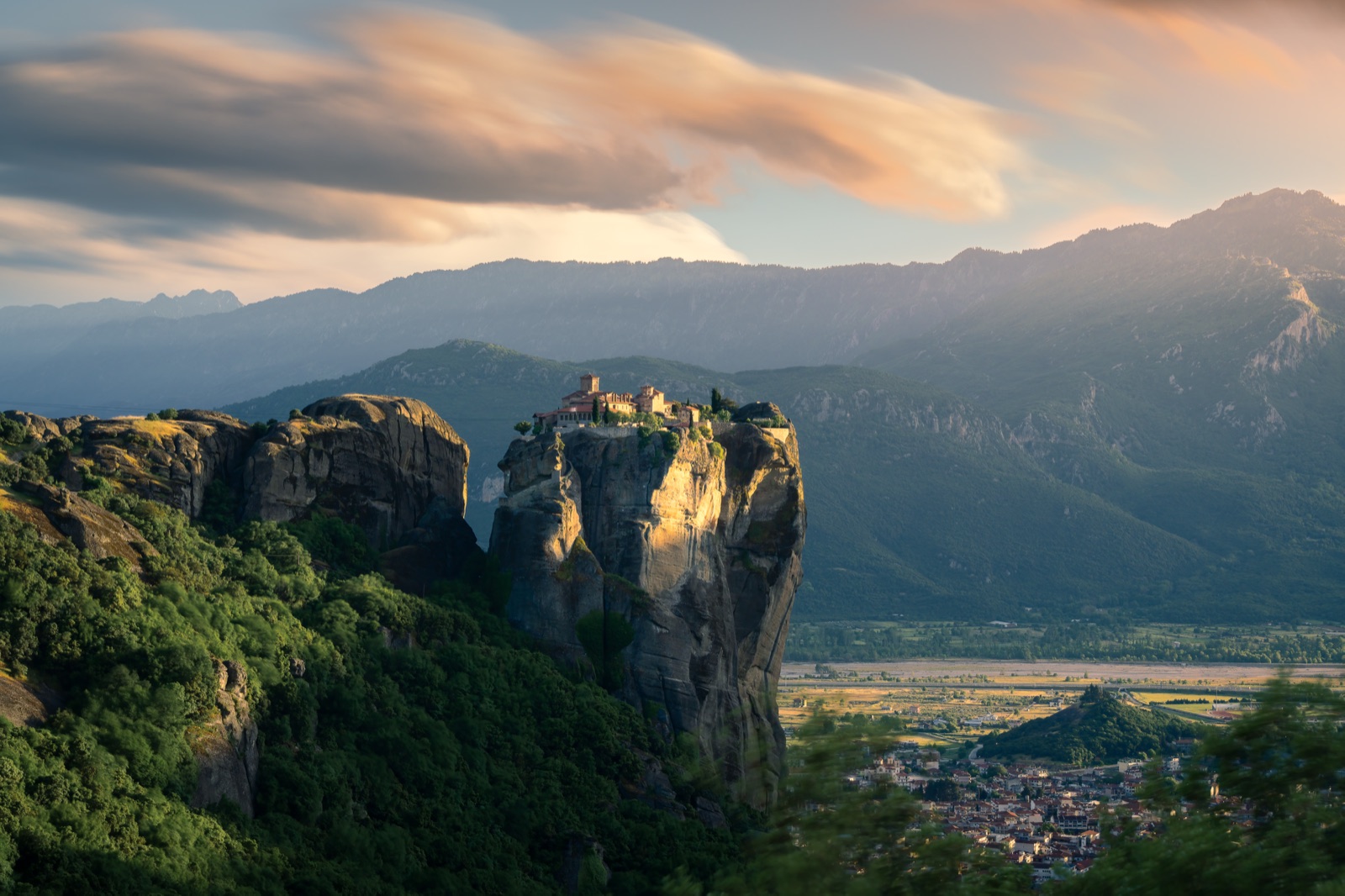 The clifftop monastery and rock pinnacles of Meteora lit warm by low evening sun, with soft pink lenticular clouds catching the last light above the mountain ridge behind