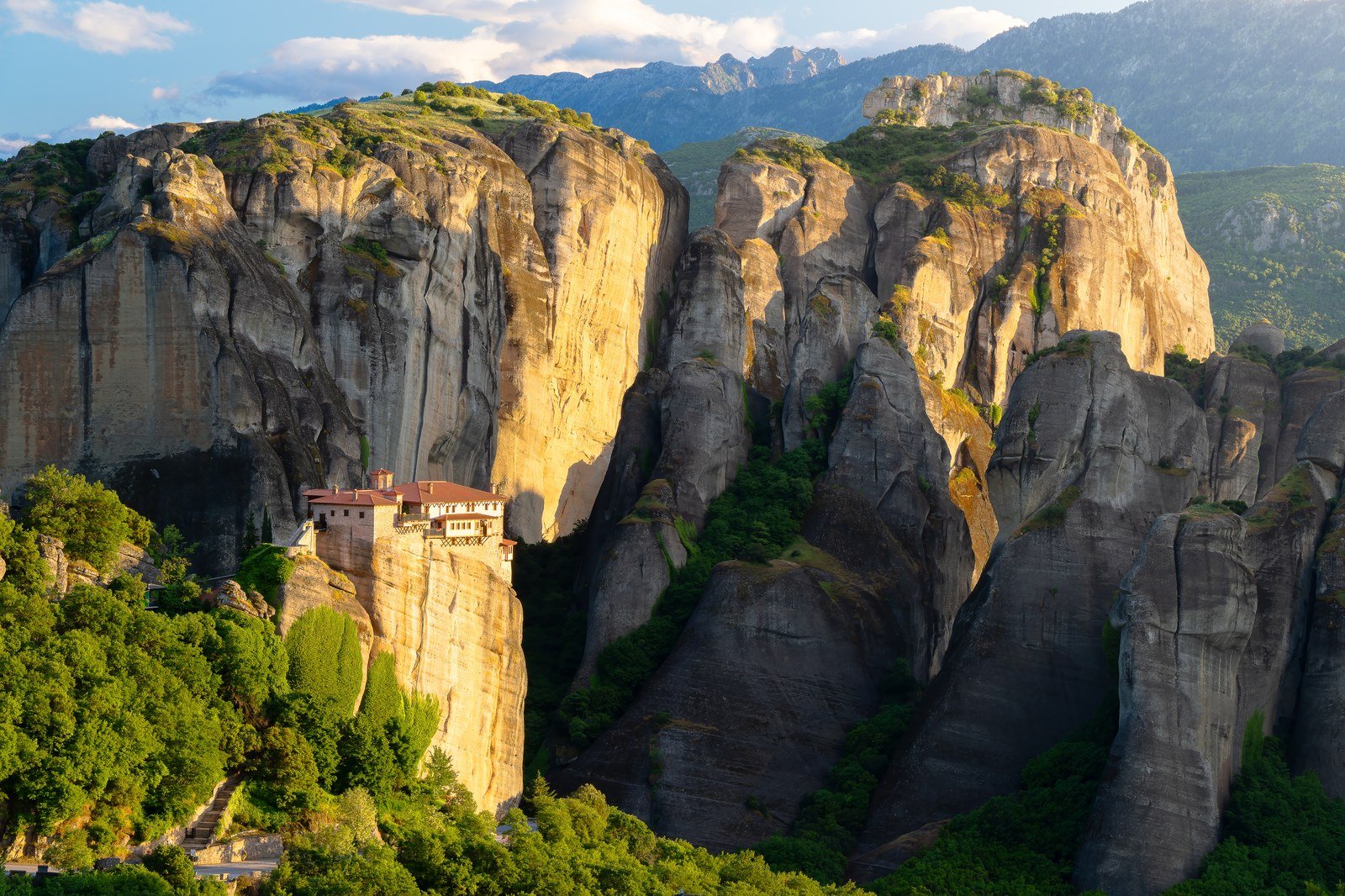A tiny clifftop monastery dwarfed by vast rock pinnacles in warm golden light, the small building giving a sense of the landscape's scale