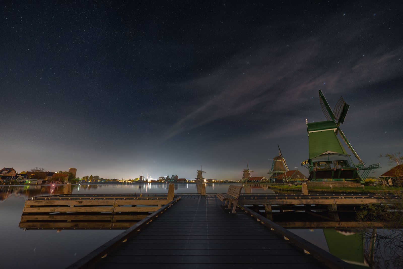 A star-filled night sky over a lamplit waterside jetty and windmills, the kind of high-ISO frame that needs careful noise reduction to stay clean without losing the stars