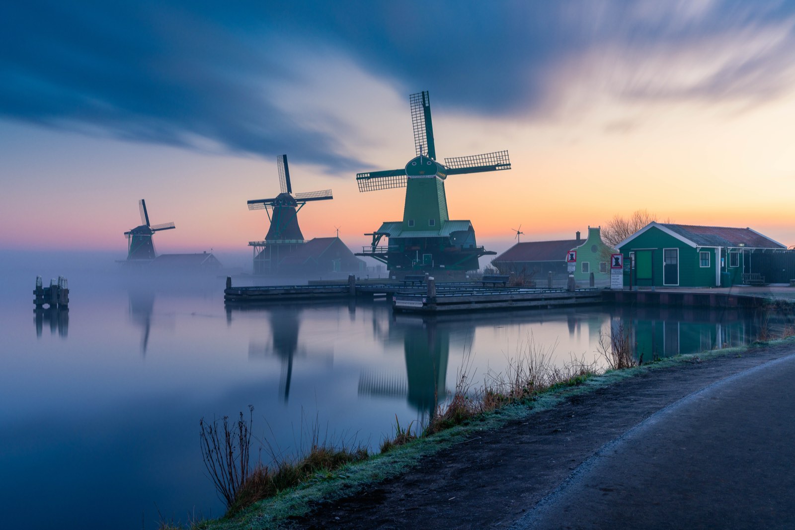 Molinos de viento históricos holandeses al amanecer reflejados en agua en calma, las nubes difuminadas en un movimiento suave por una larga exposición y una neblina baja sobre el canal