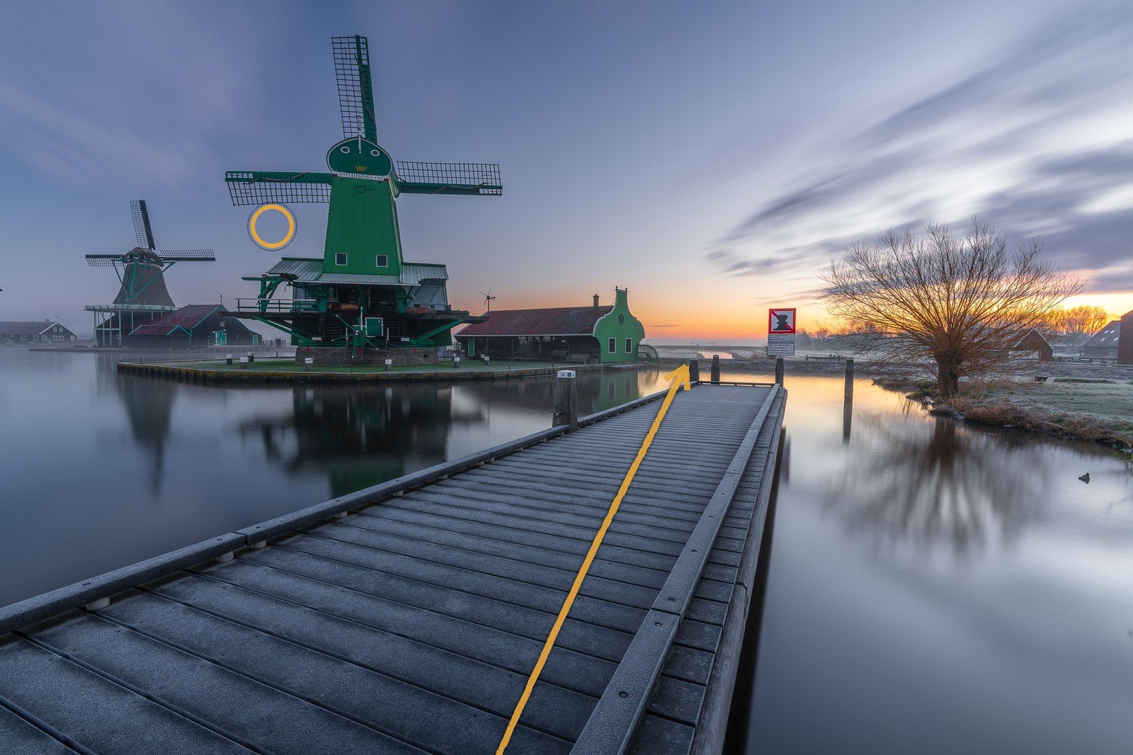 A jetty leading across still water to a green windmill at dawn, the leading line traced in amber