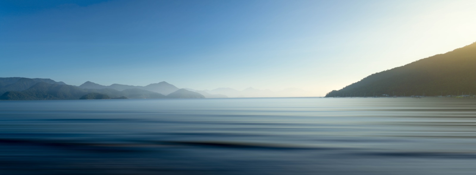 Ein Ultra-Weitwinkel-Panorama aus geschichteten blauen Bergkämmen, die im Dunst verschwinden, über einem spiegelglatten Meer bei erstem Licht in Langzeitbelichtung