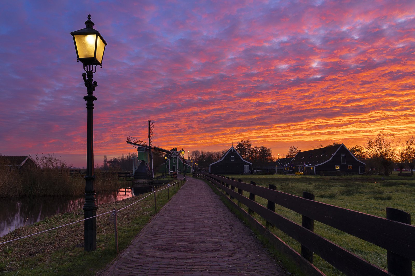 A fiery red and orange broken-cloud sky over a Dutch village with a windmill and a lit street lamp, the low sun underlighting scattered cloud into intense colour — the partial-cloud sweet spot for dramatic light