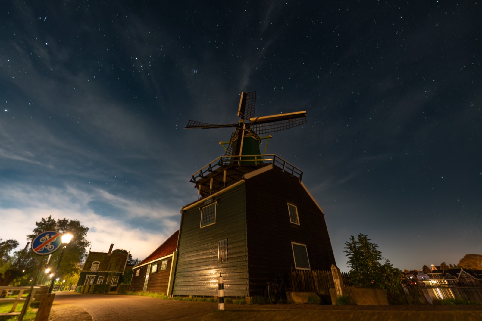 Eine historische niederländische Windmühle, beleuchtet vor einem sternenübersäten Himmel, ihre Details im Dunkeln herausgearbeitet, ein Dorf leuchtet tief am Horizont