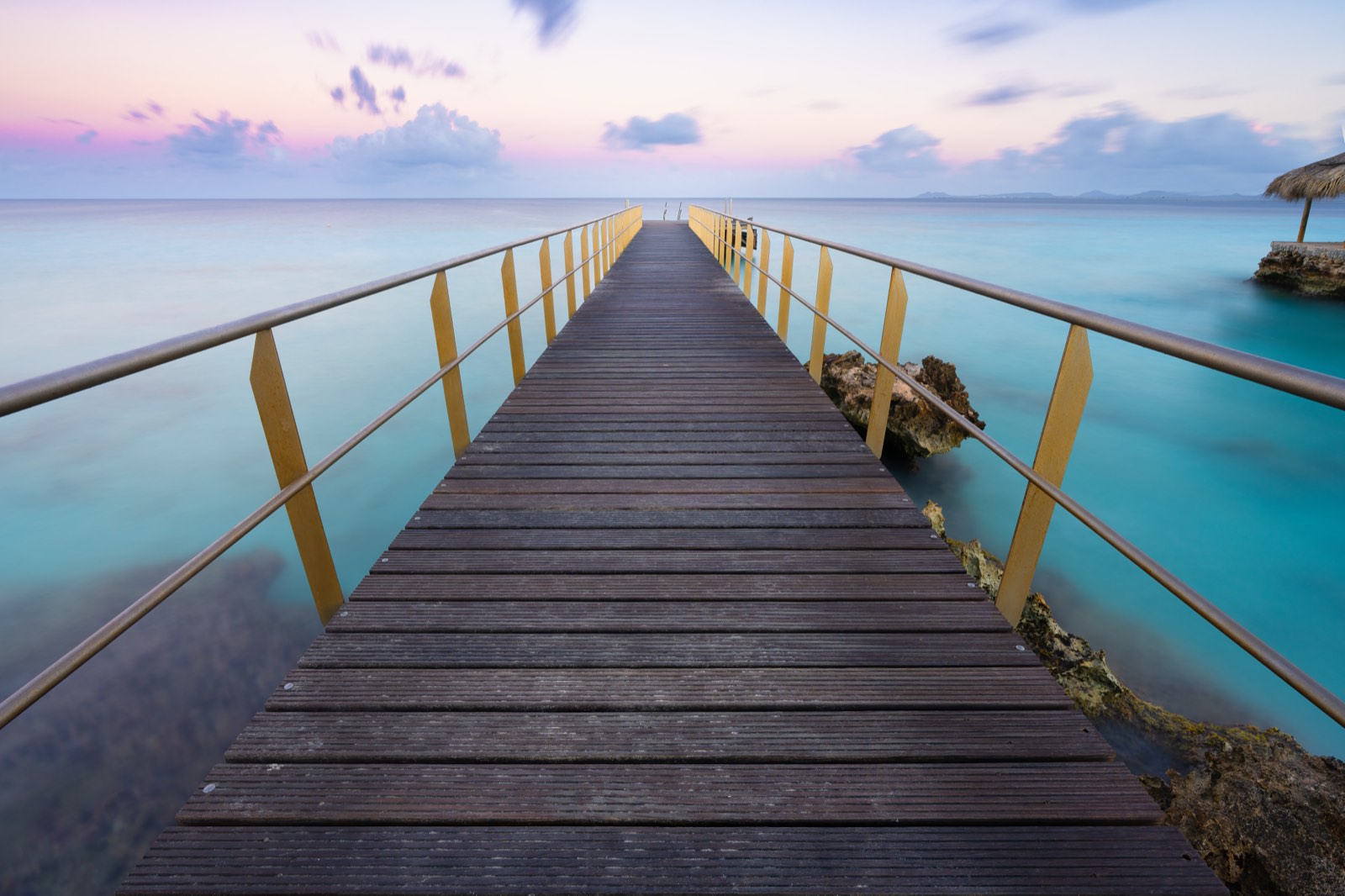 Ein hölzerner Steg, der bei Morgendämmerung über ruhiges türkisfarbenes, tropisches Wasser hinausführt, der pastellfarbene Himmel spiegelt sich im stillen Meer, eine entfernte Insel am Horizont